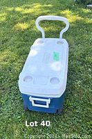 Blue and white Coleman cooler with white handle and four molded drink holders on lid, shown outdoors on grass.