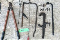 Photo showing a selection of oversized tools on a wooden surface including long-handled loppers, brace bit, coping saw, pipe clamp, and bar clamp.