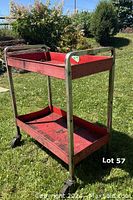 Red metal tool cart standing outdoors on grass, showing two shelves and metal handles with casters underneath.