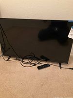 Front view of the 40-inch Vizio TV showing black bezel, the screen with some reflections, power cord plugged in, and remote control placed on the floor in front.