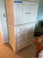 Full front view of white armoire showing upper cabinet with two paneled doors and four drawers below.