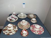 Photo showing six bone china place settings with tea cups, saucers, dessert and dinner plates in floral patterns with gold trim.