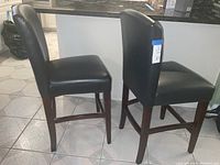 Side view of two black leather bar chairs showing straight backs, padded seats, and wooden legs with footrests.