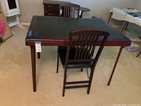 Dark wooden folding table with a faux leather top surface, two matching wooden folding chairs with padded seats placed around it, in a carpeted room.