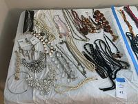 Assortment of various costume jewelry necklaces laid on a white surface, showing a variety of styles, colors, and materials including beads, faux pearls, metal floral designs, and wooden discs.