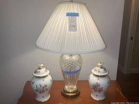 Crystal table lamp flanked by a pair of floral ginger jars on a wooden surface, showing lamp shade and bulb.
