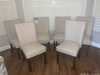Six beige fabric upholstered dining chairs arranged together on a hardwood floor next to white window shutters.