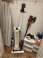 Ironing board, Dyson Cyclone vacuum, upright fan, storage drawers and accessories arranged in laundry room corner.