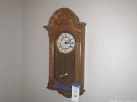 Full view of the wooden wall clock showing the curved top, clock face with Roman numerals, and brass pendulum behind glass.