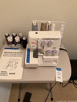 Front view of Juno Janome 3434D differential feed sewing machine on folding table with thread spools, instruction manual, and foot pedal visible.