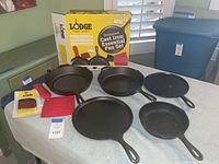 Lodge Seasoned Cast Iron Essential Pan Set in original box with visible brand and set description, includes two red silicone handle covers and silicone hot pad, two used cast iron pans in front