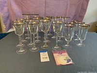 Front view of entire stemware set showing three groups of glasses with gold rims on the top edge arranged in rows, alongside a packet of glass tags and a lot number card.