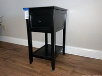 Front view of black end table showing drawer and lower shelf, tag visible.
