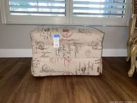 Side view of rectangular storage ottoman with travel themed fabric exterior, showing the handwritten script and postal stamp pattern, positioned on wooden floor in front of window.