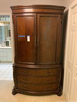 Front view of wooden TV armoire showing two closed doors and three drawers below with ornate metal handles.