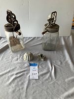 Two Dazey No 20 glass butter churns with metal lids and wooden crank handles, three vintage metal jello molds and one vintage metal cookie cutter displayed on a table.