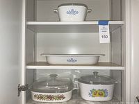Four pieces of vintage Corning Ware including small casserole dish on top shelf with blue cornflower design, flat rectangular dish in middle shelf, and two casserole dishes with glass lids on bottom shelf, one with sunflower pattern.