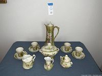 Full set of Nippon porcelain tea set arranged on table, showing teapot with lid, four cups and saucers, sugar bowl with lid, creamer, tea strainer, and small tureen with lid, all hand painted with floral designs and gold trim.