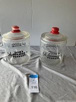Pair of clear glass jars labeled 'Eat Tom's Toasted Peanuts 5¢' with red knob lids, displayed on fabric surface.
