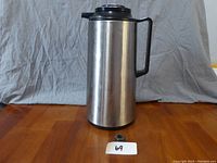 Front view of the stainless steel 1.85 litre thermos container with black handle and lid, placed on a wooden table against a neutral fabric backdrop.