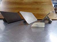Second photo, similar to first, showing three metal cooking presses on a stainless steel surface, two silver and one dark colored, all flat plates with handles.