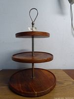 Photo showing a three-tier wooden serving stand with varying sized circular platforms and a metal rod with a loop handle on top.