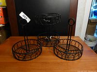 Four round black metal condiment organizers on a wooden surface. Three are flat baskets; one is a tiered, two-level basket. Each has a central vertical handle with a circular top for carrying.
