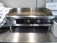 Front view of electric flat griddle showing stainless steel body, cooking plate, three control knobs, indicator lights and grease drip tray drawer.