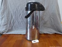 Full view of the Bunn stainless steel 2.2 litre thermos with black pump dispenser on top sitting on a wooden surface with grey backdrop.