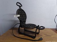 Waffle maker open with visible non-stick Belgian-style waffle plates, cord and plug on wooden table.