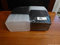 Photo showing stacks of sealed disposable plastic food containers and lids wrapped in plastic packaging on a wooden table. A label with number 191 appears on the packaging.