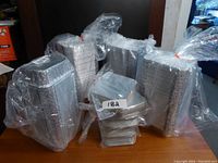 Multiple large stacks of silver foil take out trays with lids, all sealed in clear plastic packaging, arranged on a table.