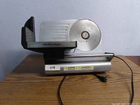 Hamilton Beach electric meat slicer with stainless steel rotary cutting blade on wooden surface against light wall background.