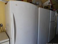 Front-left angle view of white upright freezer next to a small fridge, fan on top.