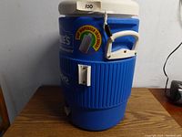 Front angle view of blue Lowe's Igloo 5-gallon cooler showing white lid, carrying handle, and side cup dispenser bracket.