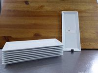 Photo showing a stack of 8 white porcelain rectangular serving platters and one platter standing upright to show shape and dimensions.