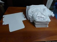 Photo showing a flat paper box sheet to be folded into a small container alongside a white plastic bag containing more boxes.