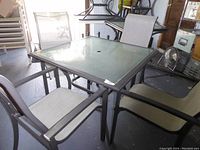 Square outdoor table with metal frame and tempered glass top with center hole, surrounded by four high-back vinyl mesh chairs.