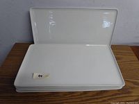 Stack of six white rectangular platters showing top and angled side view, placed on wooden surface.