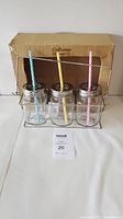 Set of 3 clear glass mason jars each with a metal lid and a striped straw (blue, yellow, pink) shown outside of the original cardboard packaging and inside a wire carrying rack.