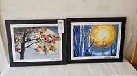Photo of two framed canvas art pictures on white surface, showing tree with pink blossoms and black cat, and blue birch tree scene with sun/moon