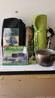 Camping gear displayed on a surface showing tents in bags, sleeping pad, camp grill box, cooking pot clustered together.