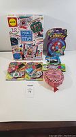 Photo showing five kids' game and activity items arranged on a white surface including the ALEX Read All About Me kit, spinner game, two Click Shots button launchers, and a pink Springy Spider ring.
