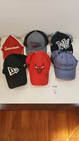Top view of six assorted baseball caps arranged in two rows. Visible brand logos include TaylorMade, Chicago Bulls, New Era, Behlen, and a stylized American flag.