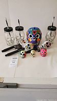 Photo of 4 clear glass skull-shaped mason jars with black lids and striped straws, one large brightly decorated ceramic skull, 3 solar-powered skull garden lights, and 4 small painted skull paperweights arranged on white surface.