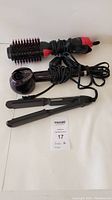 Photo showing three hair styling tools on a white surface: a round hot air brush with red bristles, a black hair straightener, and a digital cordless hair curler. All cords are coiled.