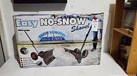 Photo of the original packaging showing the Easy No-Snow Shovel with wheels and handle on a snow background.
