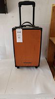 Orange portable karaoke speaker with black frame and trolley handle standing on a white surface.