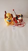 Photo showing two vintage Fisher-Price dog pull toys, one yellow with brown ears and blue collar, the other a beagle with red wheels and pull string.
