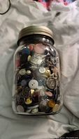 Clear glass jar filled with various buttons in different colors and shapes, showing front and side views of the jar.